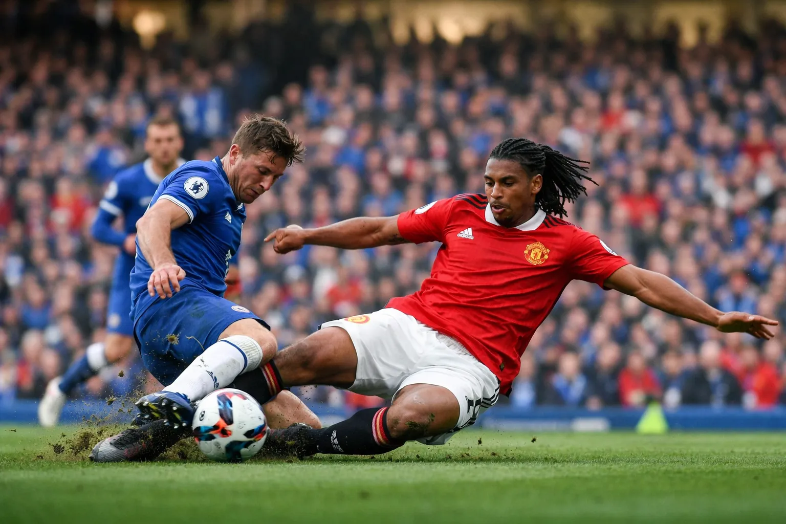 A young defender in a Manchester United kit making a sliding tackle on a Chelsea player at Stamford Bridge.