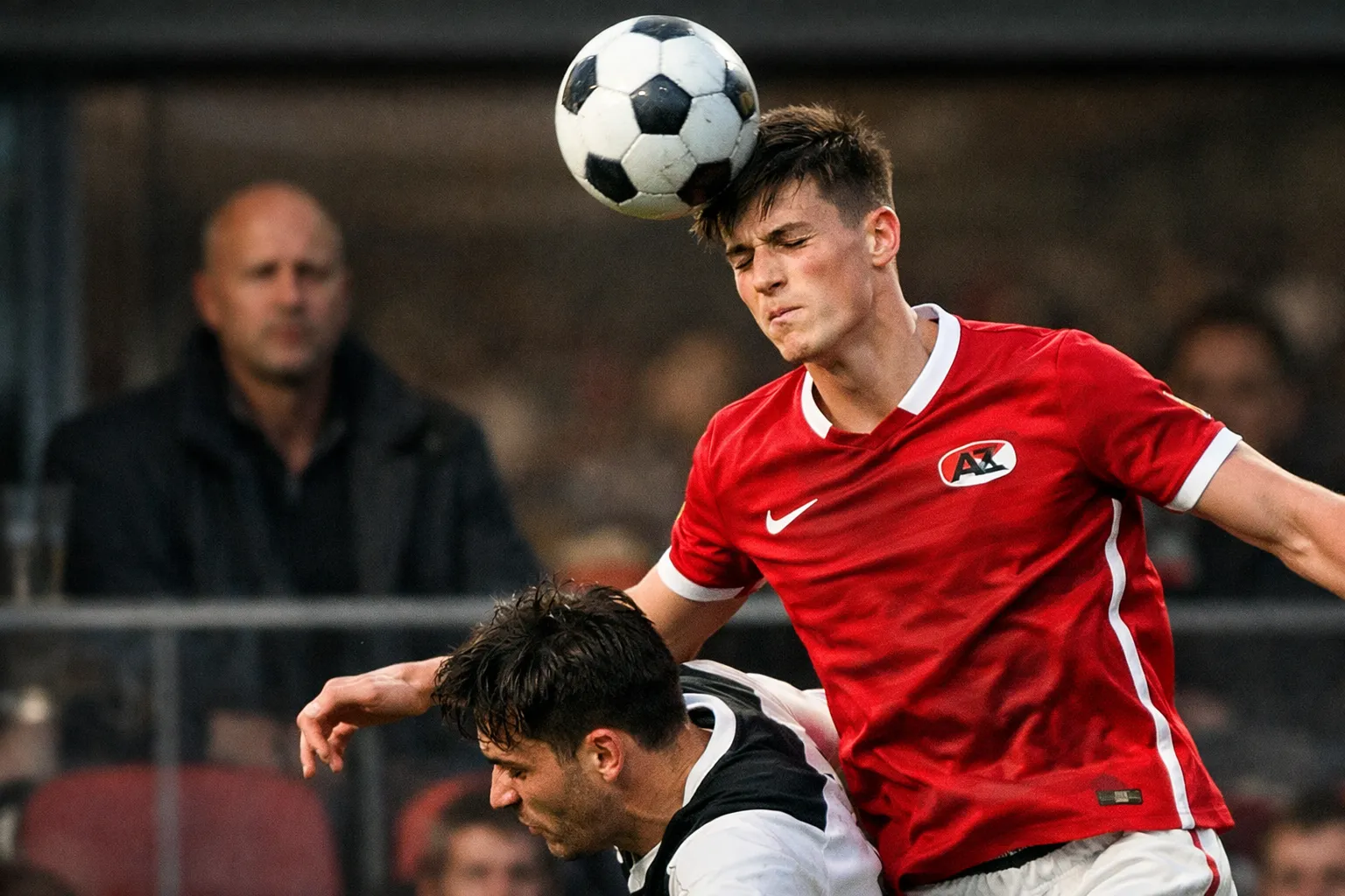 A young defender in an AZ Alkmaar kit winning a header, with a blurred figure watching from the stands.