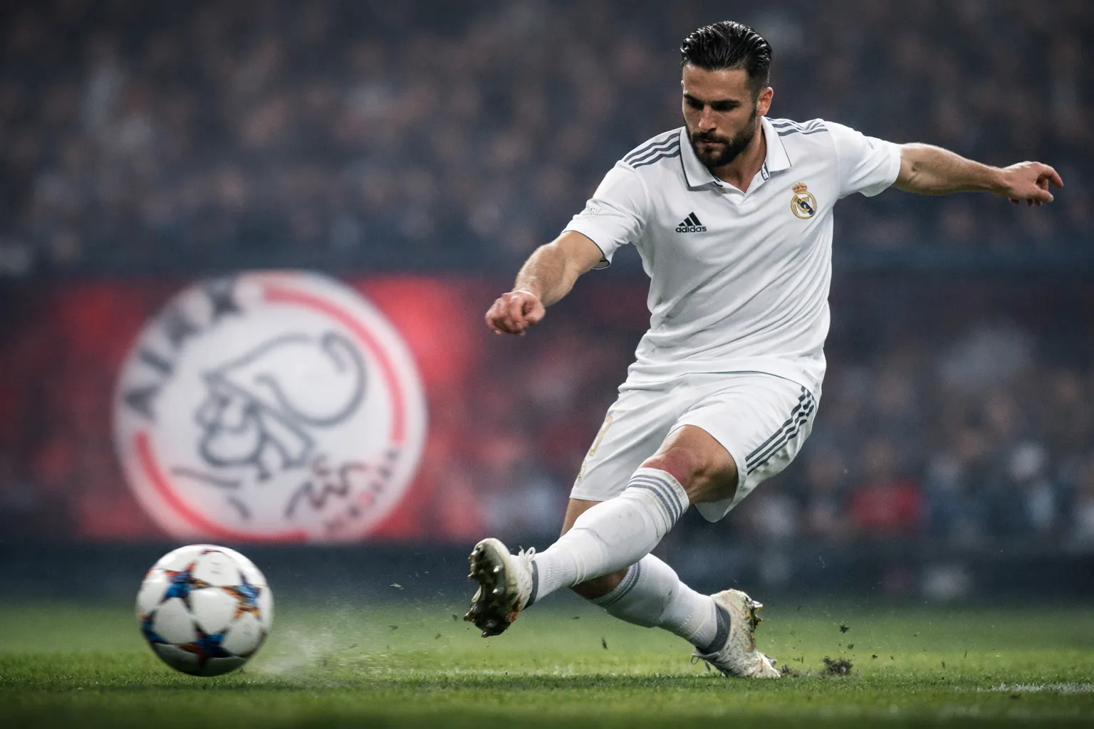 A technical midfielder in a Real Madrid kit playing a through ball, with a blurred Ajax crest on an advertising board.
