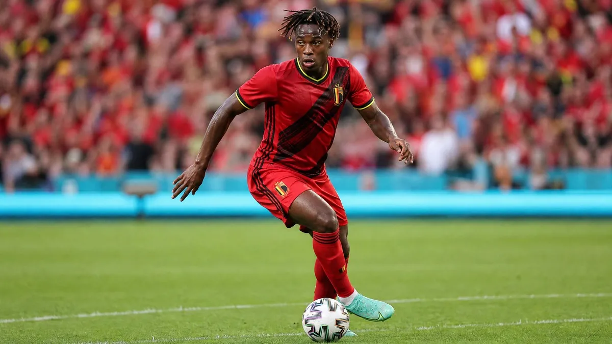 Jeremy Doku in red Belgium national team kit, mid-dribble on football pitch, focused expression, blurred stadium crowd background.