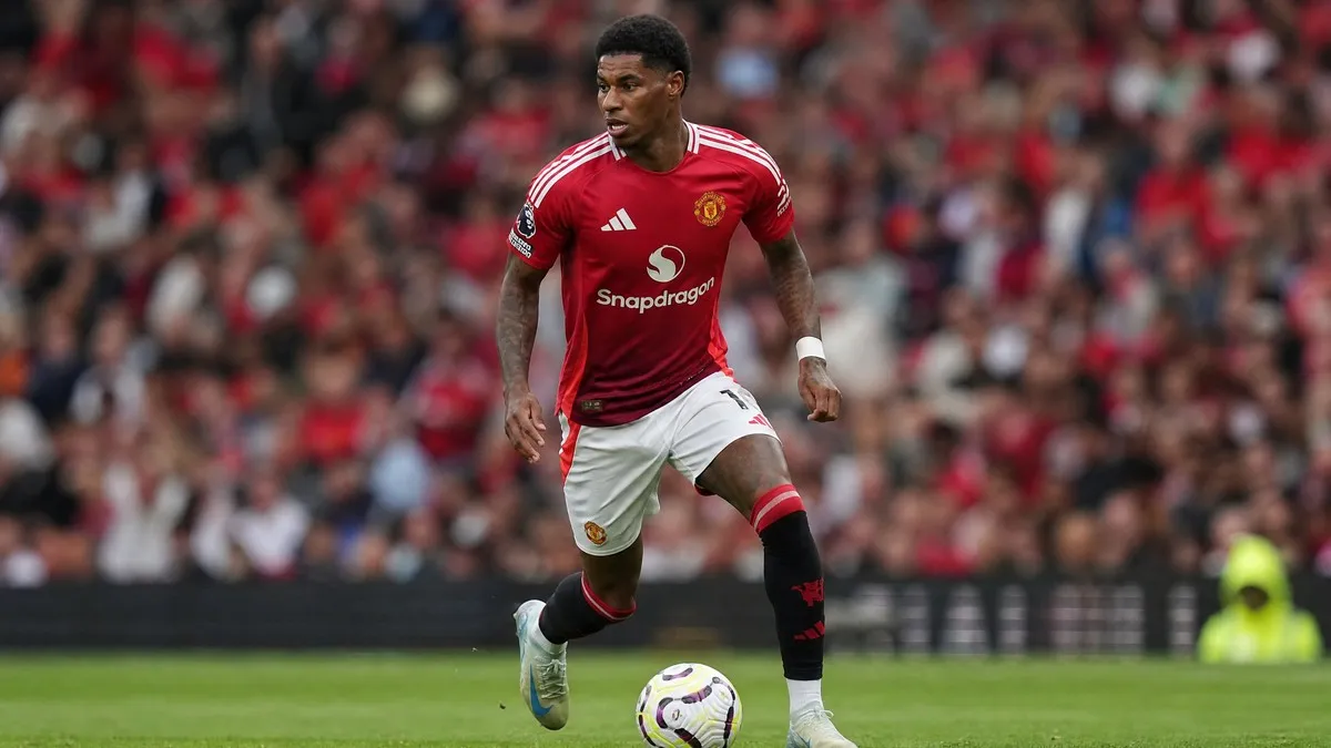 Marcus Rashford dribbling a football in his red Manchester United home kit, with a blurred stadium crowd in the background.